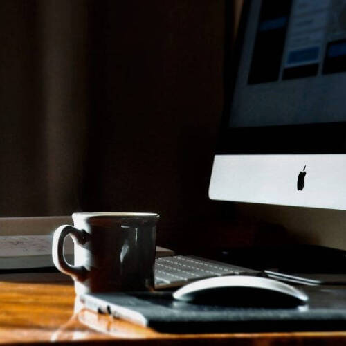 A desktop workspace featuring an Apple iMac on the right, partially visible with a white keyboard and Magic Mouse on a wooden desk, next to a dark coffee mug. Soft lighting highlights the glossy surface of the monitor and the warm wood grain of the desk.